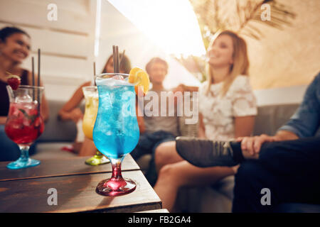 Glasses of cocktail on table with young people sitting on sofa on roof top. Friends having a rooftop party. Stock Photo