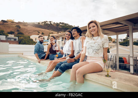 Smiling young woman looking at camera with her friends sitting on the edge of swimming pool. Young people enjoying rooftop party Stock Photo