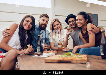 Group of friends gathered around the table at a roof party. Multiracial young people looking at camera and smiling. Stock Photo