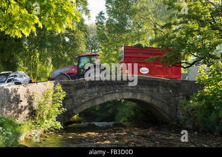 Tractor pulling a red trailer drives over an old, rustic, stone bridge crossing Clapham Beck - Clapham village, North Yorkshire, England, GB, UK. Stock Photo