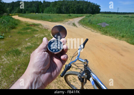 The hand of a man holding a magnetic compass on the background of the Bicycle wheel and summer trails outside of town. Stock Photo
