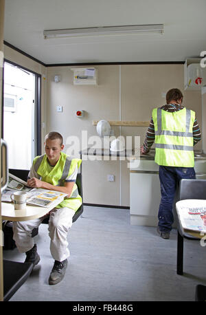 Two builders in high-vis jackets relax in a construction site cabin, making coffee and reading the paper. Stock Photo