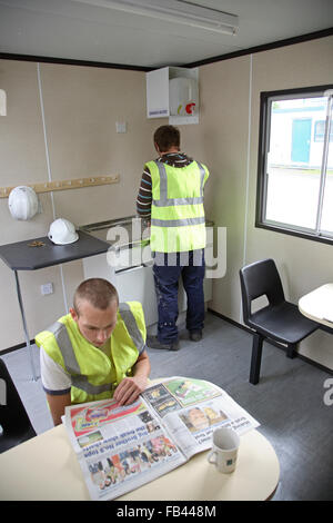 Two builders in high-vis jackets relax in a construction site cabin, making coffee and reading the paper. Stock Photo