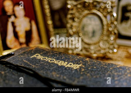 An old, well-worn album of photographs sits on a table in front of gold framed photos. Stock Photo
