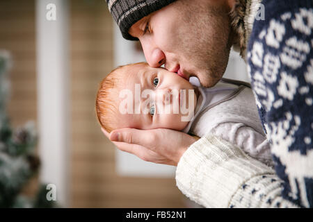 Father holding and kissing baby son among while wearing winter hat, with Christmas tree in background Stock Photo