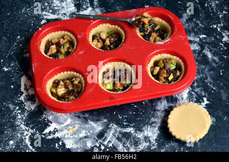 Red silicone muffin tray with unbaked open apple and mince pies on a dark floured surface Stock Photo
