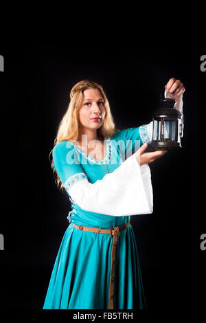 Studio shot of young girl in a medieval turquoise dress looking at the camera while holding up a retro style lamp (portrait on b Stock Photo