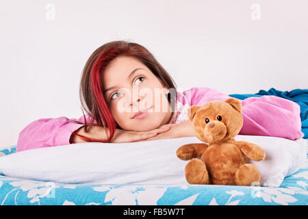 Beautiful young woman in pink pajamas lying in bed with her cute plush teddy bear, thinking of her dreams or a loved one. Stock Photo