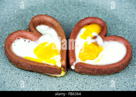 Couple of eggs cooking with heart-shaped sausages in frying pan. Cooking fried egg in the form of heart. Stock Photo