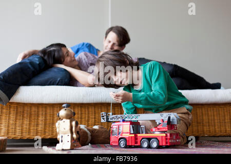 Boy playing with toys, mother and father lying on sofa in background Stock Photo
