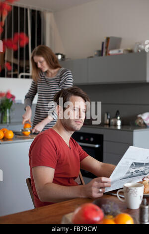 Man reading newspaper in kitchen, woman making orange juice in background Stock Photo