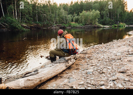 Rear view of hiker sitting on dead tree trunk at lakeshore Stock Photo