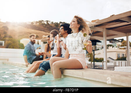 Portrait of young people sitting on the edge of the swimming pool with their feet in water. Friends relaxing by the pool during Stock Photo