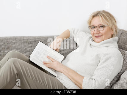 an older blonde woman lying on the sofa and reading a book Stock Photo