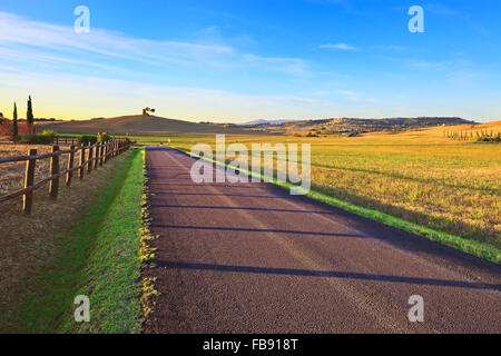 Tuscany, Maremma typical countryside sunset landscape with rural road, fence, green field, trees, rural tower and village on bac Stock Photo