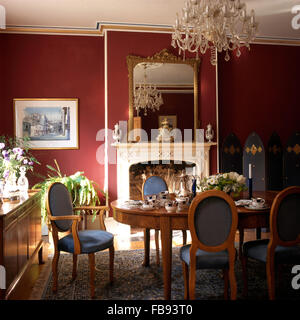 Venetian glass chandelier above antique table with blue seated chairs in a red dining room with a gilt mirror above fireplace Stock Photo