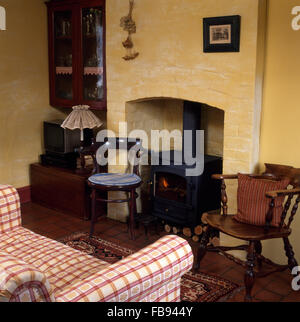 Wooden chairs either side of fireplace with wood burning stove in cottage sitting room with a red checked sofa Stock Photo