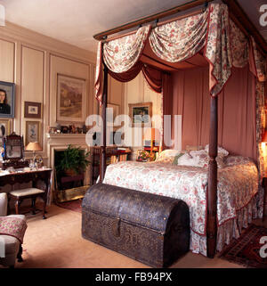 Antique studded chest below four poster bed with floral drapes in a country bedroom Stock Photo