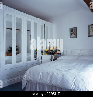 Mirrored panels in doors of fitted wardrobe in an all white bedroom with a white duvet on the bed Stock Photo