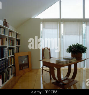 Thirties style table and fitted bookcase in a modern loft conversion study with white blinds on floor to ceiling windows Stock Photo
