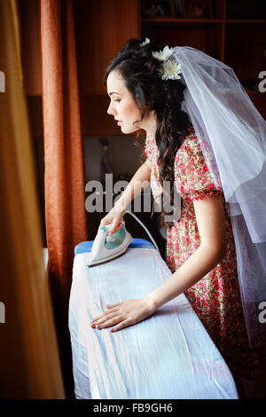 The bride preparing for wedding ceremony, ironing her gown on board, natural light. She is dressed in a colorful retro dress looking out window thinking about future. Stock Photo