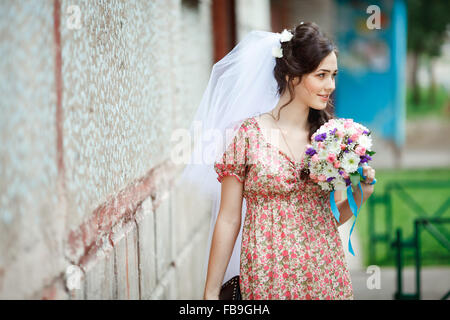 The bride in simple retro dress with floral pattern, already wearing veil, wedding bouquet and handbag, posing outside house, looking to side. Stock Photo