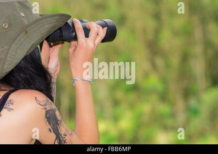 Brunette wearing green safari hat and tattoos on shoulder, holding up binoculars  looking forward, forest background, shot from behind angle Stock Photo