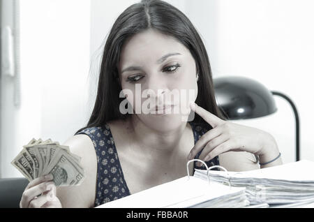 Hispanic brunette office woman sitting by desk with paper files archive open and holding several ten dollar bills. Stock Photo