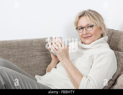 an older blonde woman lying on the sofa and drinking a cup of tea Stock Photo