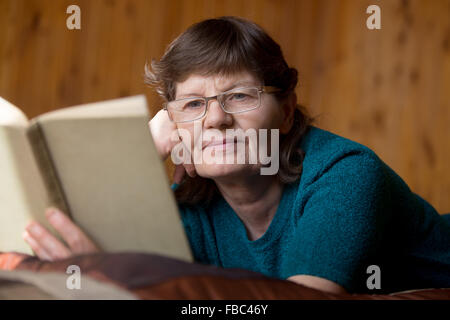 Beautiful elder woman in spectacles reading a book with faint smile, lying relaxed on bed at home in comfy bedroom with wooden w Stock Photo