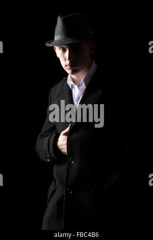 Handsome mysterious Caucasian man in hat and coat standing in shadow, low key studio shot against black background Stock Photo