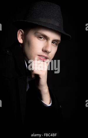 Portrait of smiling good-looking young man in hat and coat with confident look, studio shot against black background Stock Photo