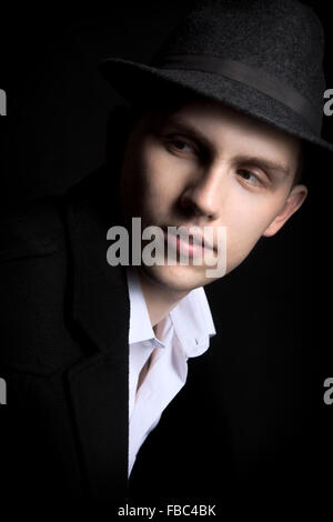 Low key shot of young man in dark hat and coat on black background, looking away from camera Stock Photo