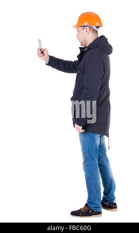 Backview of an engineer in construction helmet stands and using a mobile phone. Standing young guy. Rear view people collection. backside view of person. Isolated over white background. Foreman photographs on the phone what he sees. Stock Photo