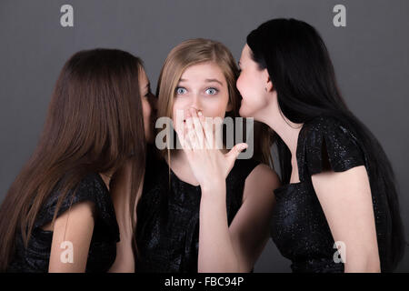 Three young girlfriends gossiping, teenage girl looks shocked as two her friends are murmuring rumors in her ears Stock Photo