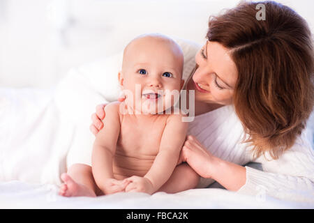 Mother and child on a white bed. Mom and baby boy in diaper playing in sunny bedroom. Parent and little kid relaxing at home Stock Photo