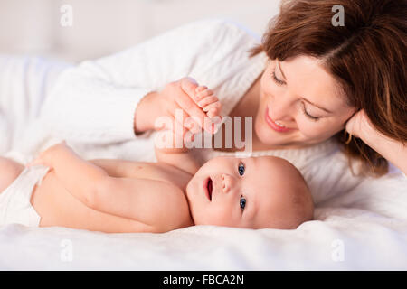 Mother and child on a white bed. Mom and baby boy in diaper playing in sunny bedroom. Parent and little kid relaxing at home Stock Photo