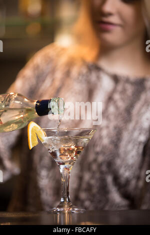 Blond female bartender preparing alcohol drink at bar counter, holding bottle, pouring drink in cocktail glass, close-up, focus Stock Photo