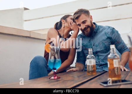 Two young people sitting together gossiping. Woman whispering something in man's ears during a party. Stock Photo