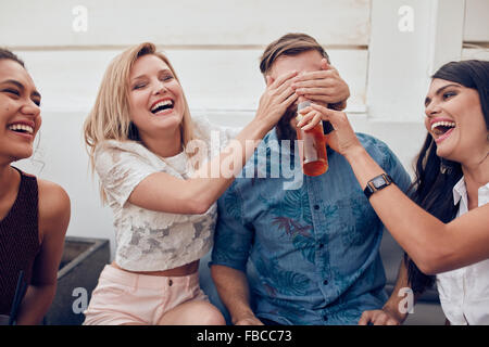 Shot of young people sitting together enjoying party. Woman closing eyes of a man with another giving drink. Young friends havin Stock Photo