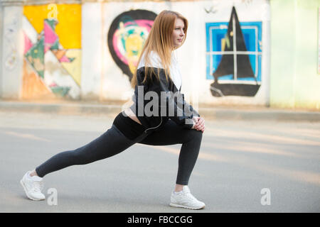 Runner girl with earphones in sportswear doing lunge exercises before morning jogging on the sunny street in front of wall with Stock Photo