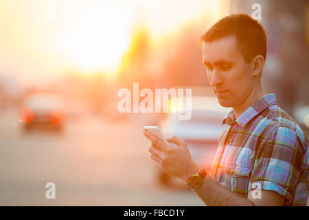 Young man holding mobile phone, using smartphone app, scrolling, looking at screen, standing on sunny street with transport vehi Stock Photo