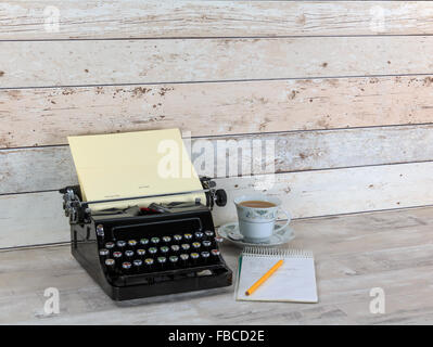 Vintage typewriter with notepad, pencil and china cup filled with tea on an old shabby wood effect desk and background Stock Photo