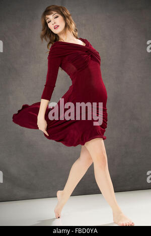 Attractive young woman dancer in long burgundy tango dress, turning with dress swirling, on gray background. Stock Photo