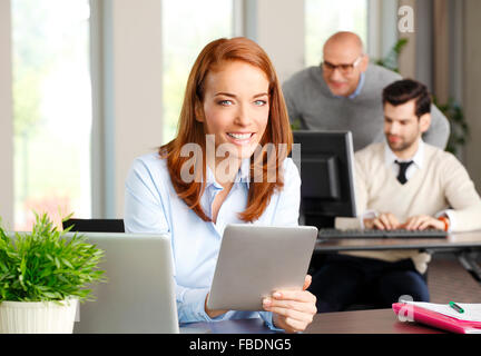 Portrait of attractive business woman sitting at desk in front of computer and analyzing financial data on digital tablet. Stock Photo