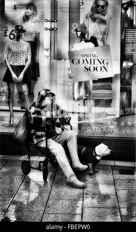 Elderly lady sitting on her shopping trolley outdoors next to shop window.  Dog at her feet Stock Photo