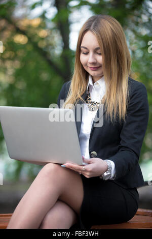 Smiling girl in elegant classic outfit using laptop, study, working, communicating, buying on-line, sitting on the bench in city Stock Photo