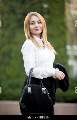 Portrait of young smiling woman on summer street dressed in formal elegant outfit: black skirt and white shirt, carrying her jac Stock Photo