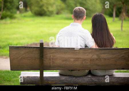 Couple of young lovers sitting close on wooden bench in park on a date, back view Stock Photo