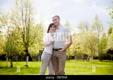 Portrait of loving couple in casual clothes, standing together in garden, smiling, young man embracing his girlfriend Stock Photo